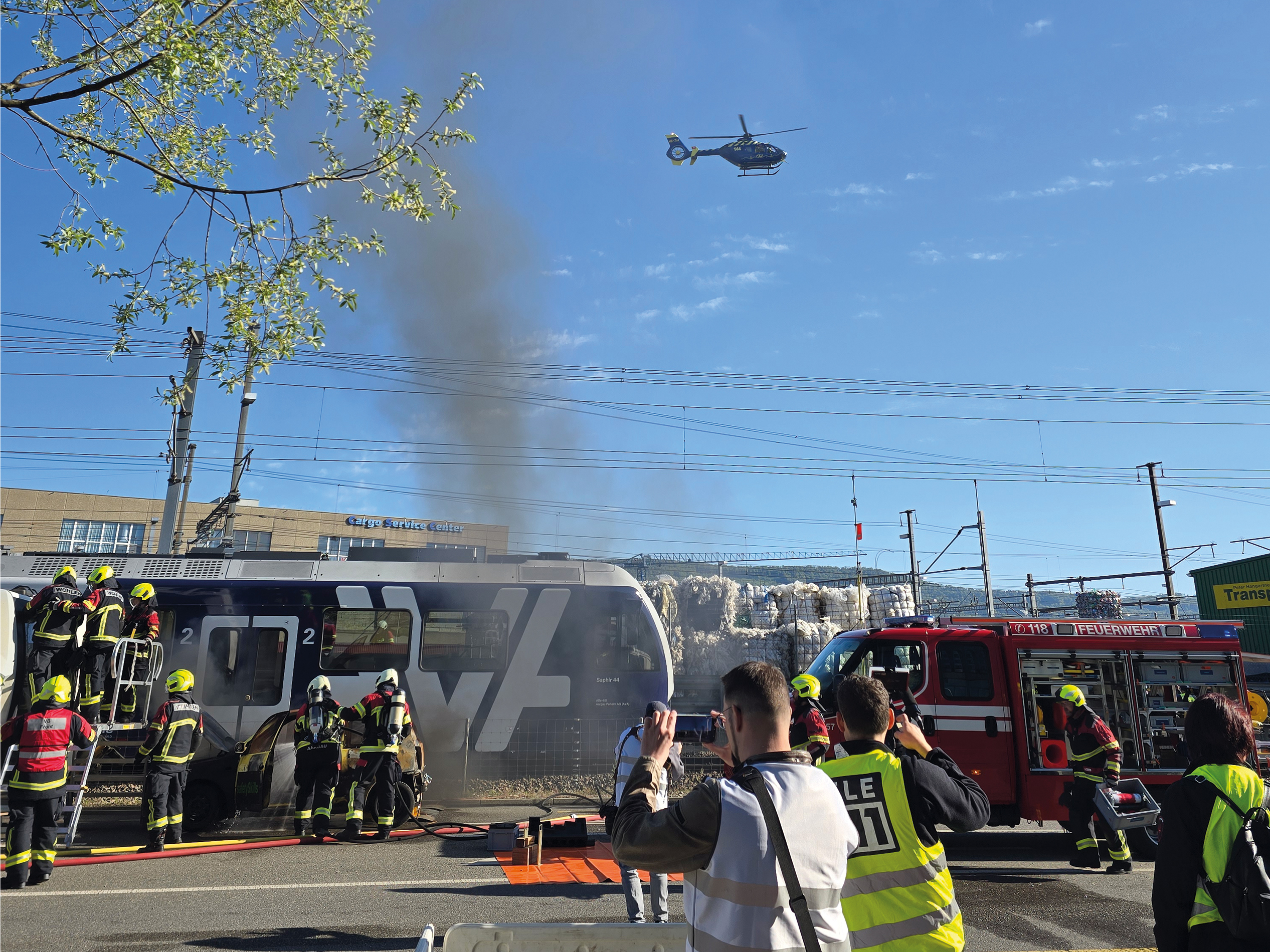 Momentaufnahme aus der Chaosphase: Die Feuerwehr verschafft sich Zugang zum hinteren Waggon, der AAA-Rettungshelikopter überfliegt die Szenerie, während Beobachter alles dokumentieren.